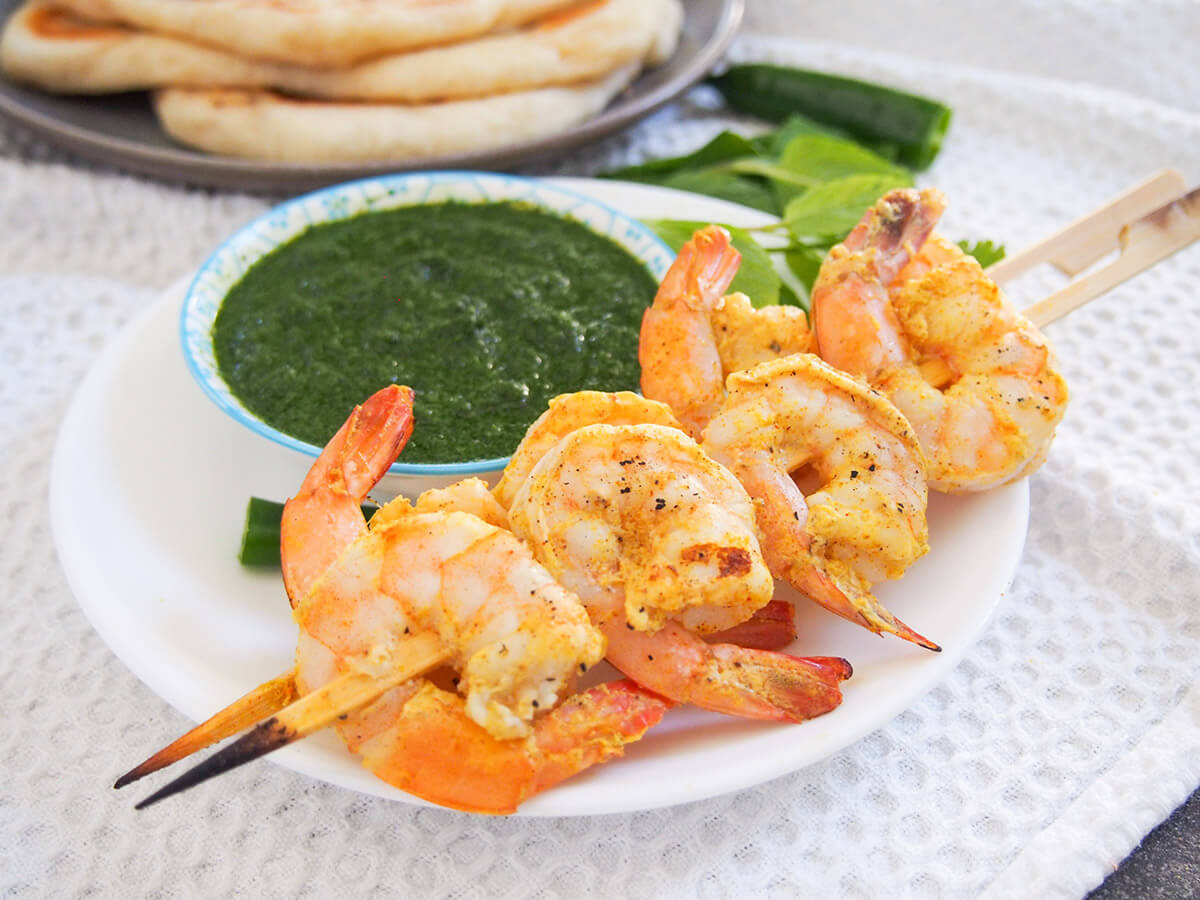 Tandoori shrimp are marinated in a spiced yogurt mixture then grilled to give a delicious flavor. Great served simply with naan bread and chutneys and/or raita. two skewers of tandoori shrimp with mint chutney behind and naan in background