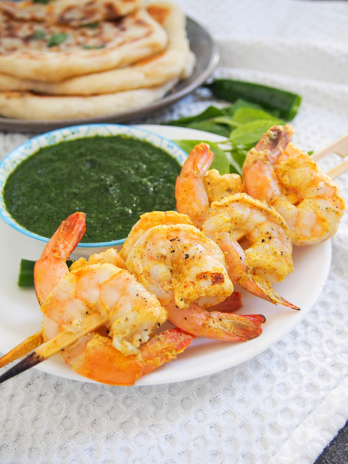 Tandoori shrimp are marinated in a spiced yogurt mixture then grilled to give a delicious flavor. Great served simply with naan bread and chutneys and/or raita. two skewers of tandoori shrimp with chutney behind and plate with naan in background