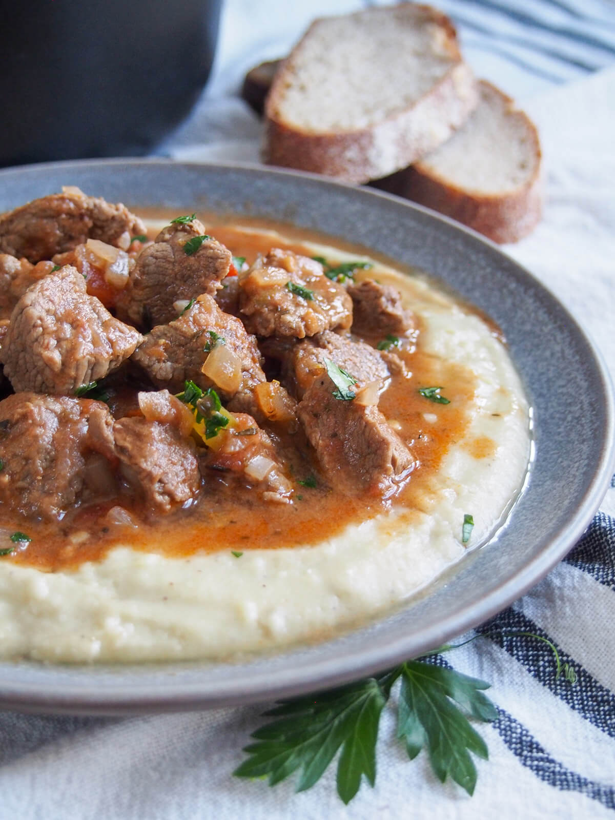 side view of plate of lamb stew over eggplant-bechamel sauce with pieces of bread behind plate
