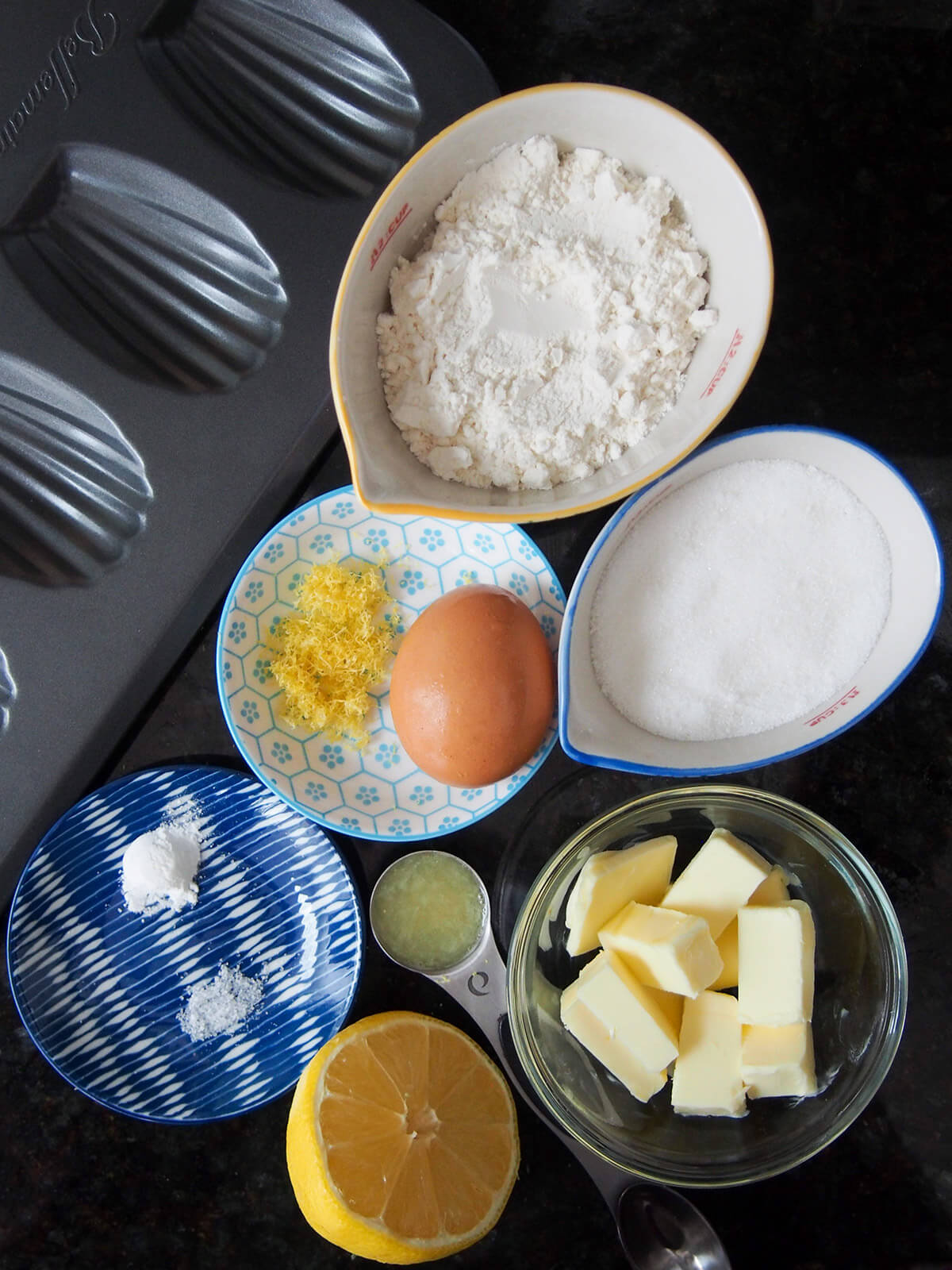 ingredients for lemon madeleines dishes with flour, sugar, butter, egg, lemon and moulded pan to side