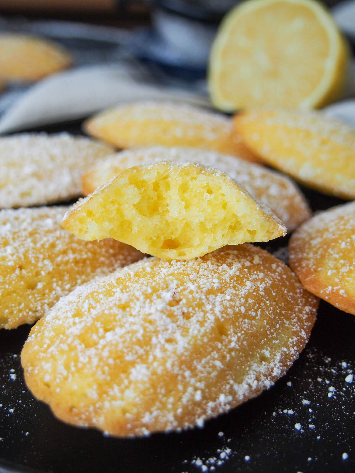 Lemon madeleines are a classic French treat. This small batch recipe makes the perfect amount to enjoy the light, sweet and gently crisp deliciousness at its best. a bitten into madeleine sitting on top of others with lemon behind