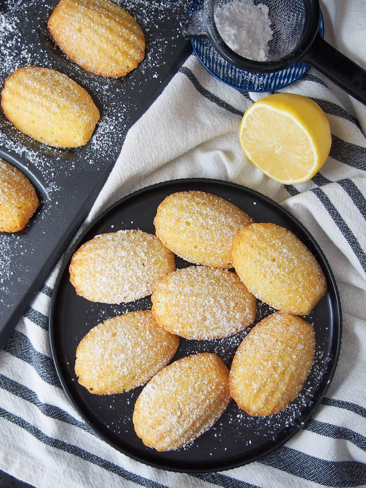 Lemon madeleines are a classic French treat. This small batch recipe makes the perfect amount to enjoy the light, sweet and gently crisp deliciousness at its best. plate of lemon madeleines from overhead with cut lemon and sifter with sugar above