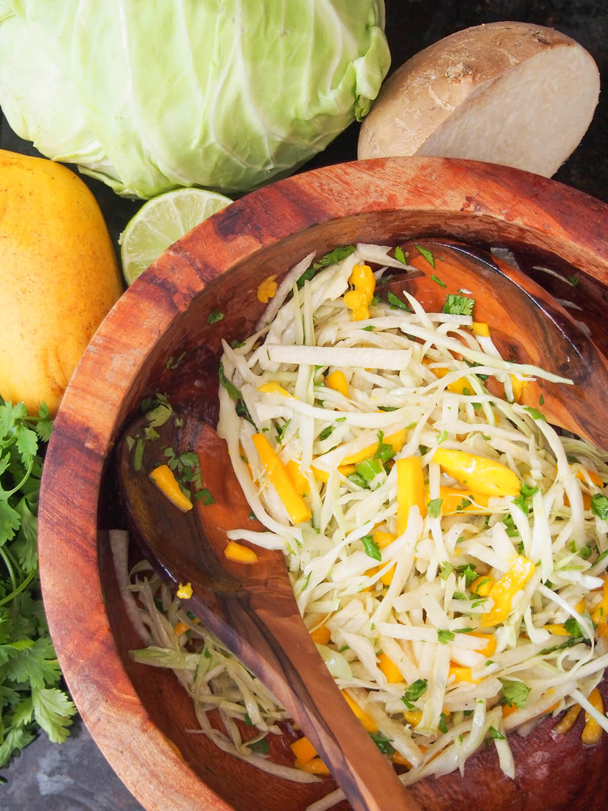 wooden bowl with mango jicama slaw and wooden salad servers around it to mix and ingredients to side of bowl