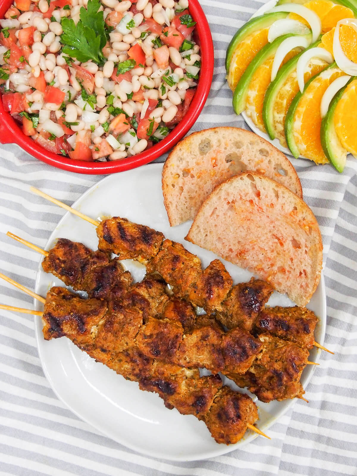Pinchos morunos are Spanish pork skewers with an aromatic spice marinade. They're easy to make, full of flavor and the perfect tapas dish to add to your next party or get-together. overhead view of plate of pinchos morunos and pan con tomate with plates of salads above