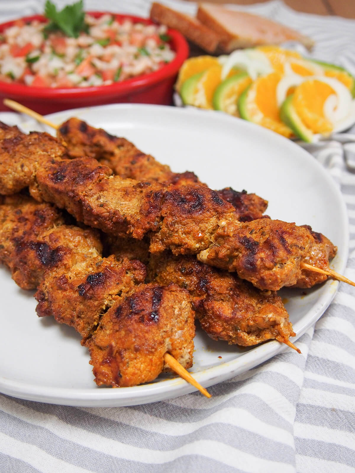 Pinchos morunos are Spanish pork skewers with an aromatic spice marinade. They're easy to make, full of flavor and the perfect tapas dish to add to your next party or get-together. white plate with pinchos morunos with salads behind