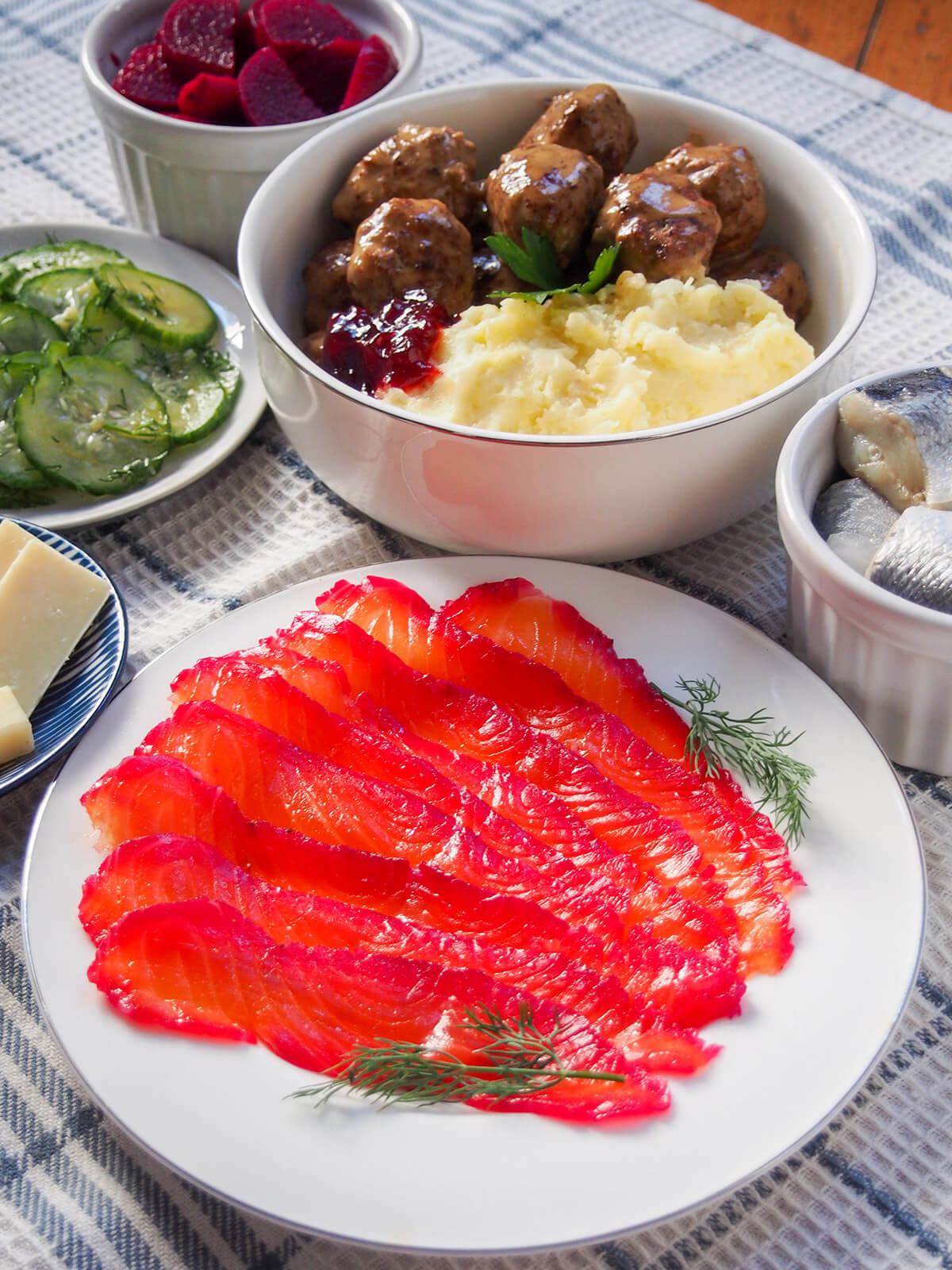 This beet cured salmon is a simple twist on gravlax where the salmon takes on a beautiful purple hue on the one side. Yes, you have a little wait, but it's easy to make and the result is delicious and perfect for elegant appetizers. plate of beet cured salmon with dishes of meatballs and potato, pickled beets and cucumbers behind