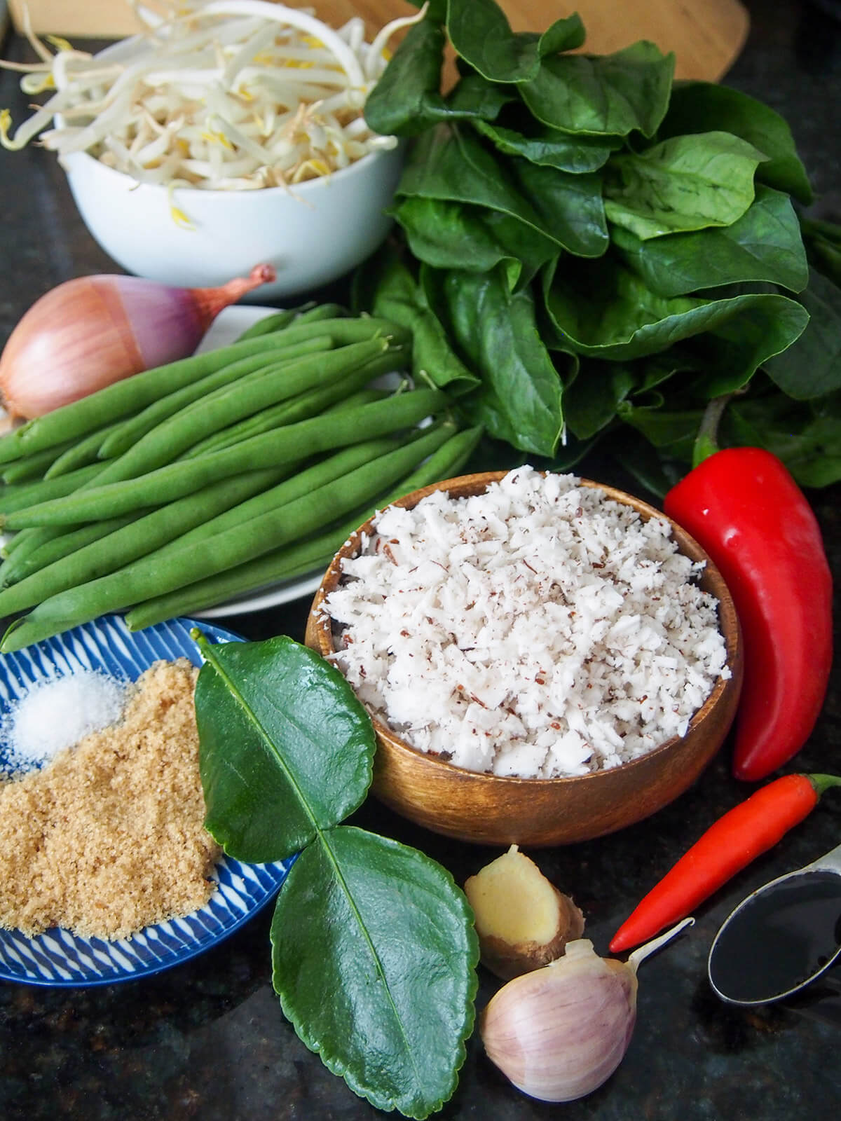 Ingredients for urap sayur - Indonesian vegetables with coconut bowls with coconut, beans, beansprouts with chilis, spinach, lime leaves and sugar to side