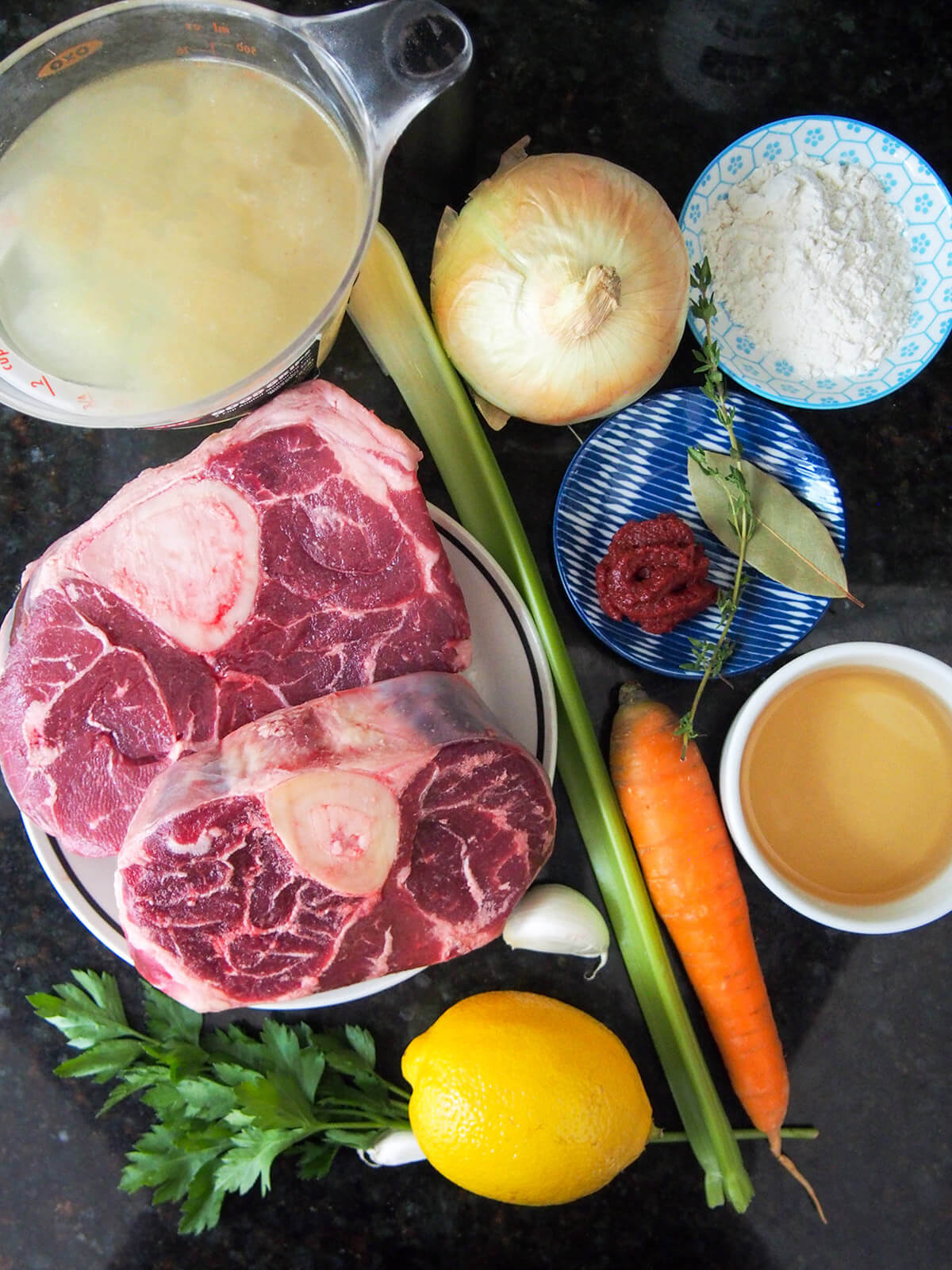 ingredients for beef osso bucco dishes with beef shank, stock, wine, four, tomato paste, herbs, onion and veg to side