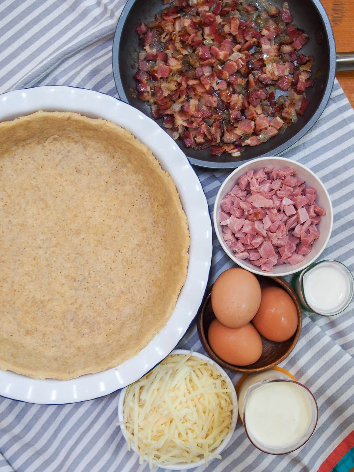pie case, skillet with bacon and onion, bowls with cheese, eggs, cream and ham