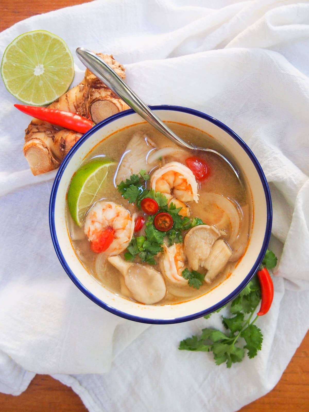 overhead view of bowl of tom yum soup with lime galangal one side, cilantro chili other