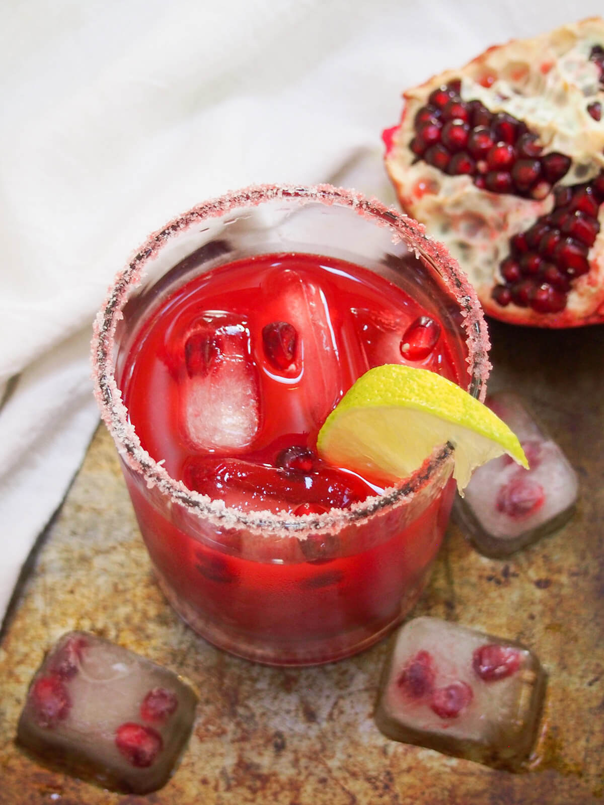 glass of pomegranate margarita with ice cubes below and pomegranate behind