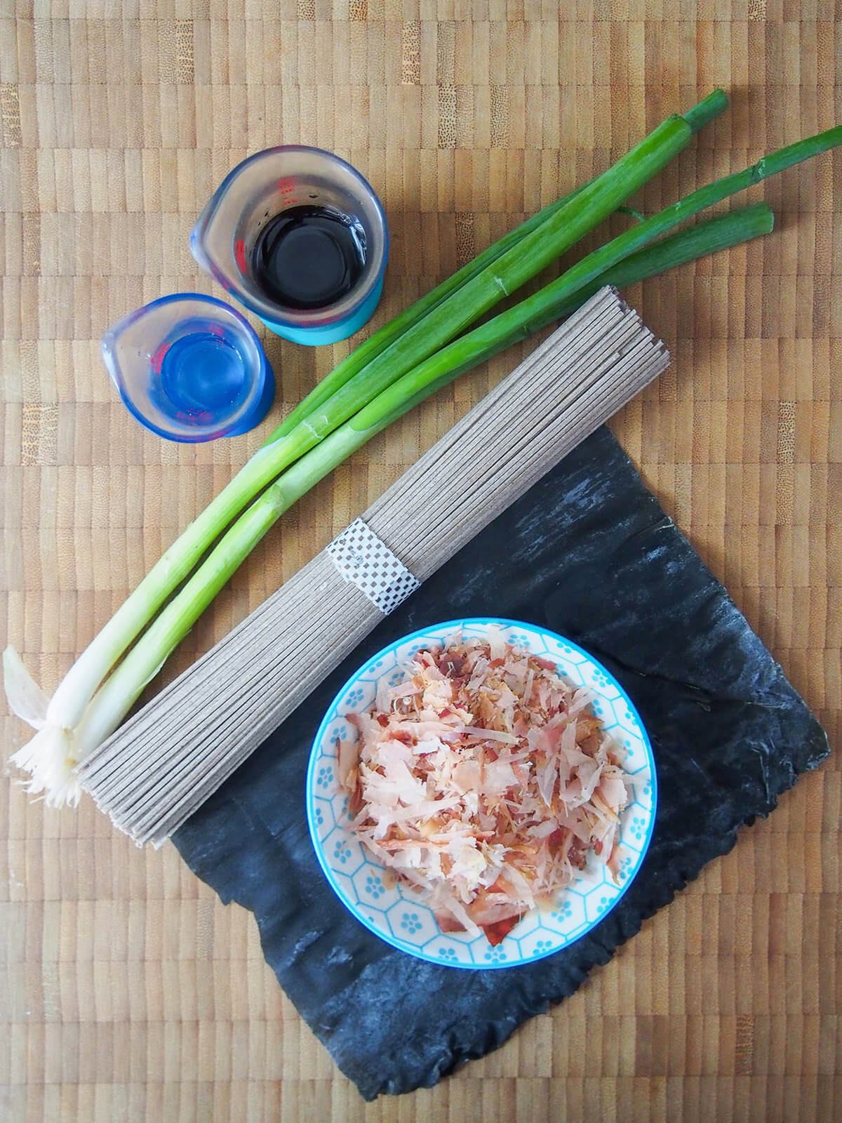 green onions, bundle of noodles and dish with bonito flakes on top of piece of kombu seaweed
