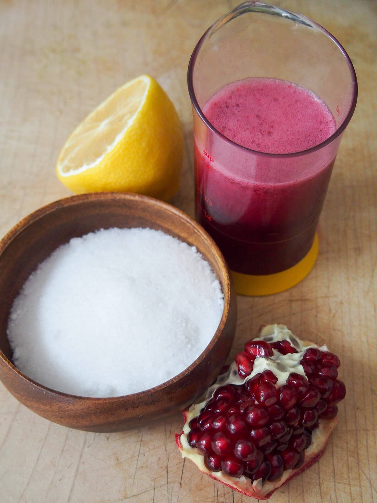 bowl of sugar, piece of pomegranate, beaker with pomegranate juice and half lemon