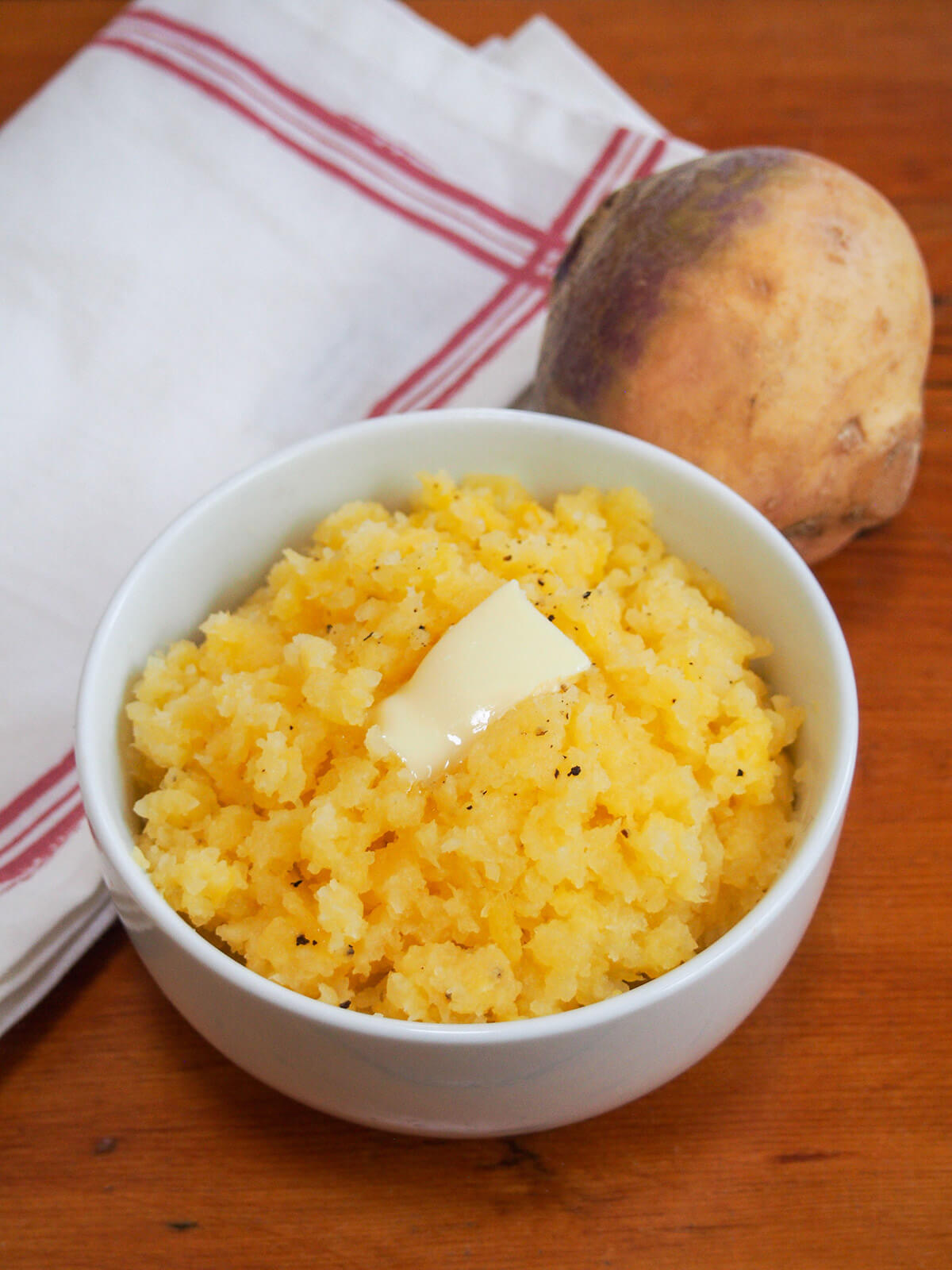 Mashed rutabaga, or swede mash, is an easy side dish and tasty alternative to mashed potato or other root vegetables. It pairs perfectly with a range of mains like roast meats and sausages with a mild and gently sweet flavor. bowl of mashed rutabaga with rutabaga (or swede) and cloth behind