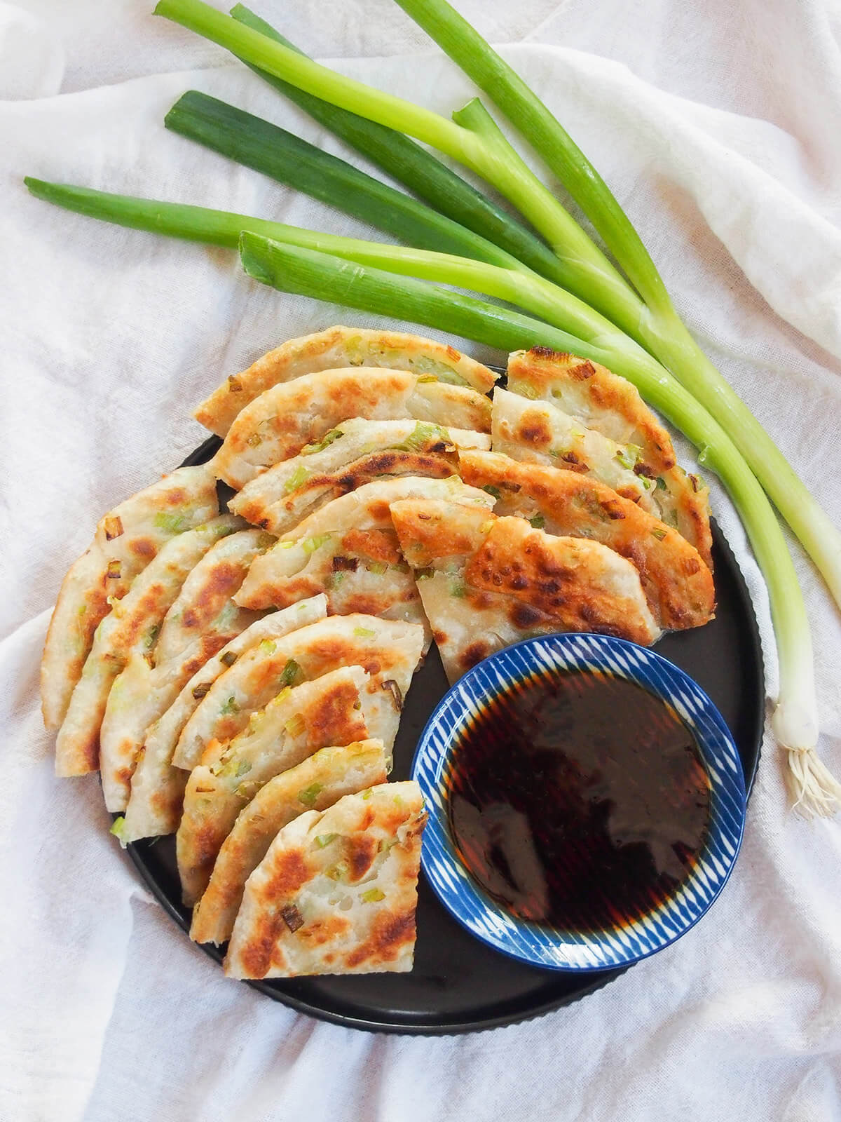 Chinese scallion pancakes are a street food and restaurant favorite. They're easier than you might think with a delicious mix of flaky layers, chewiness, crisp outside and tasty flavors. plate of Chinese scallion pancakes (cong you bing) from overhead with scallions above plate