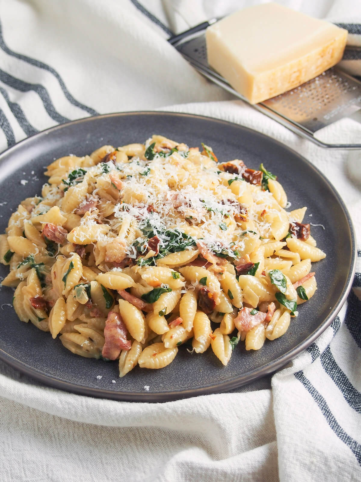 plate of bacon sun dried tomato pasta with spinach and cream with block of parmesan behind