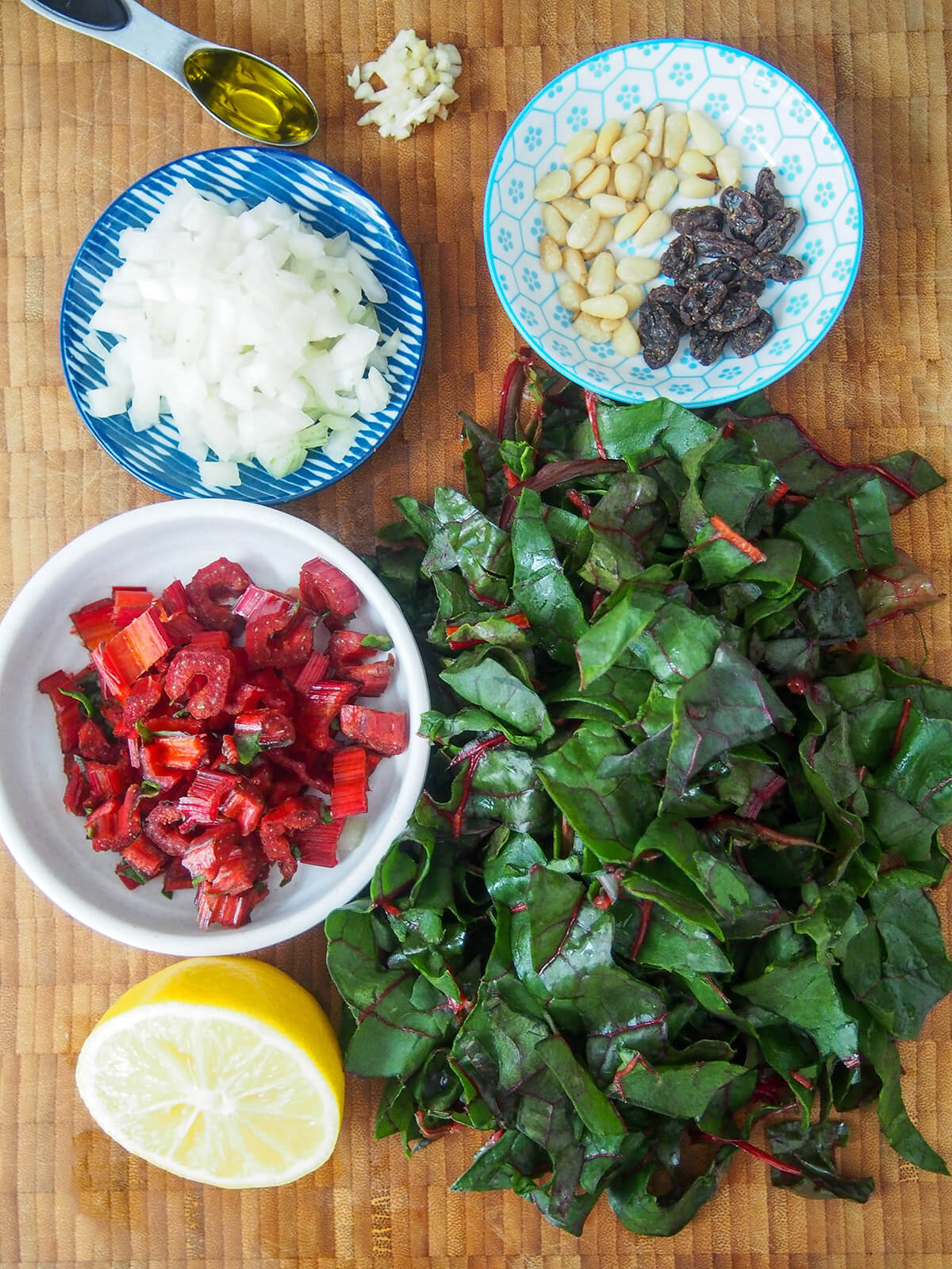 Ingredients for sauteed chard with pine nuts and raisins stack of chard leaves with plates with onion, pine nuts, raisins, chard stems to side and lemon