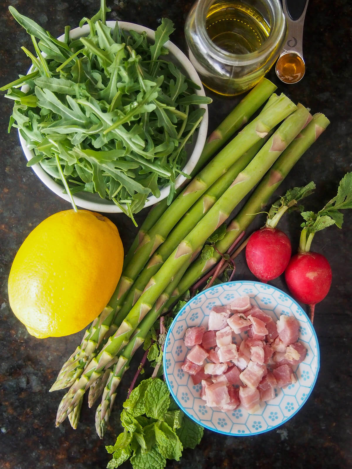 bowls with arugula, pancetta and asparagus, lemon, radishes and some mint and jar with oil in between