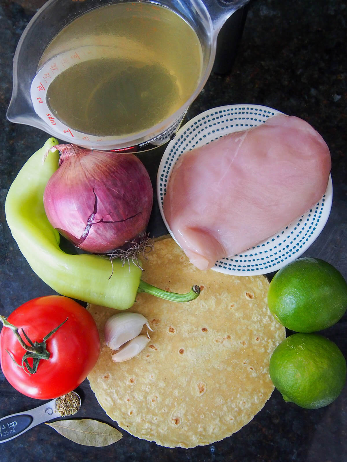 chicken breast on plate next to limes, red onion, pepper, tomato, tortilla and stock