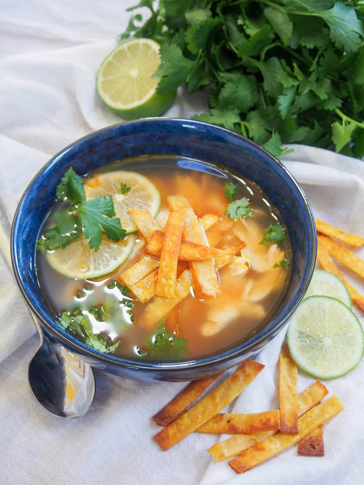bowl of sopa de lima with spoon on one side, bunch of cilantro behind and lime and tortilla strips on other side