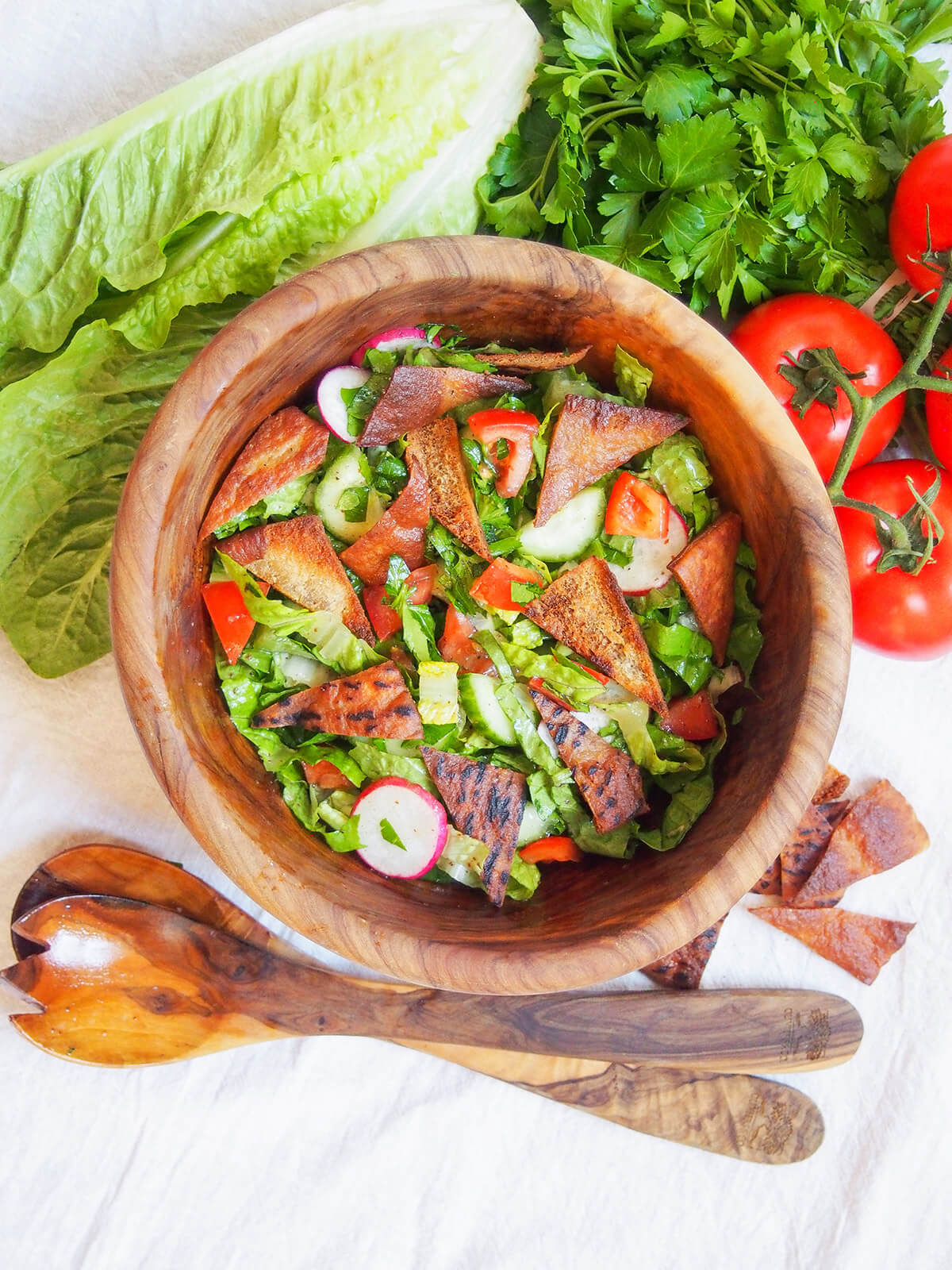 bowl of fattoush salad with lettuce, parsley and tomatoes above bowl and serving spoons below