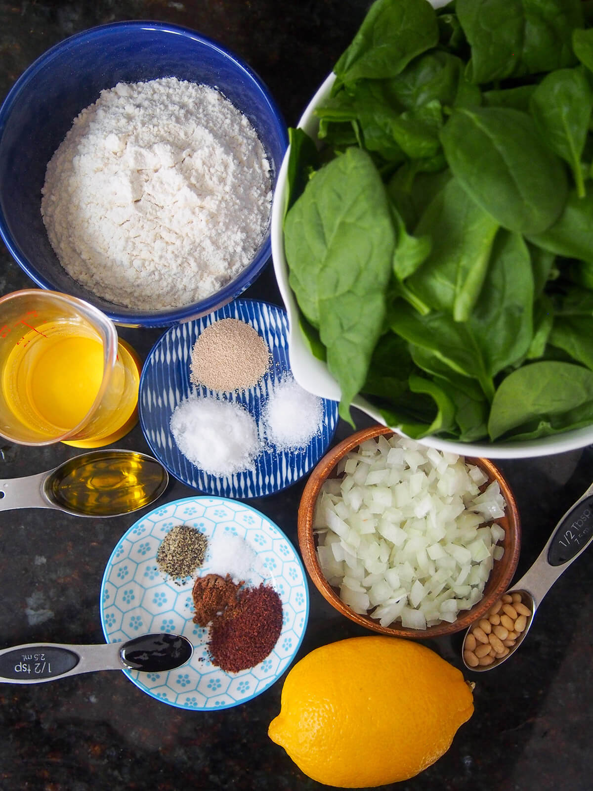 bowls with spinach, flour, onion and small plates with spices, salt and yeast with lemon, water and oil to side