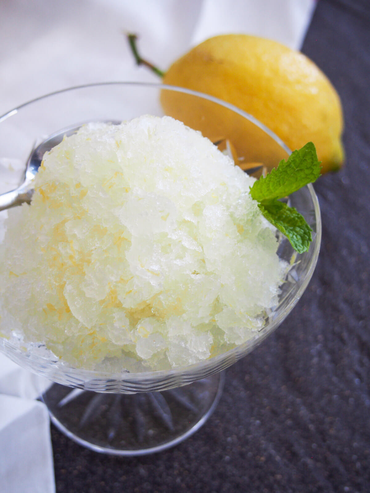 part view of glass of lemon granita from side with lemon behind and spoon in back of glass