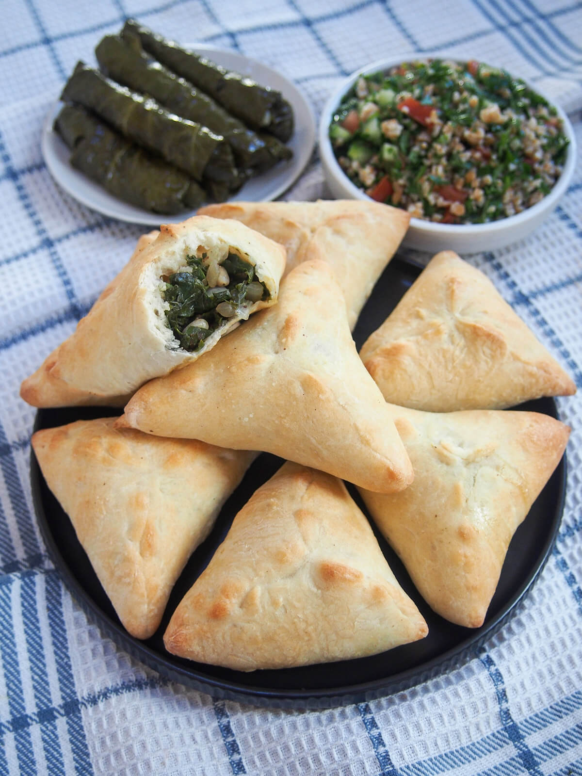 bitten into spinach fatayer resting on top of plate with more any stuffed grape leaves and tabbouli behind