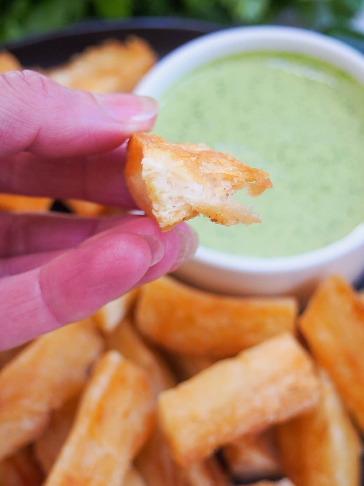 holding a bitten yuca fry in hand with blurred fries and sauce behind