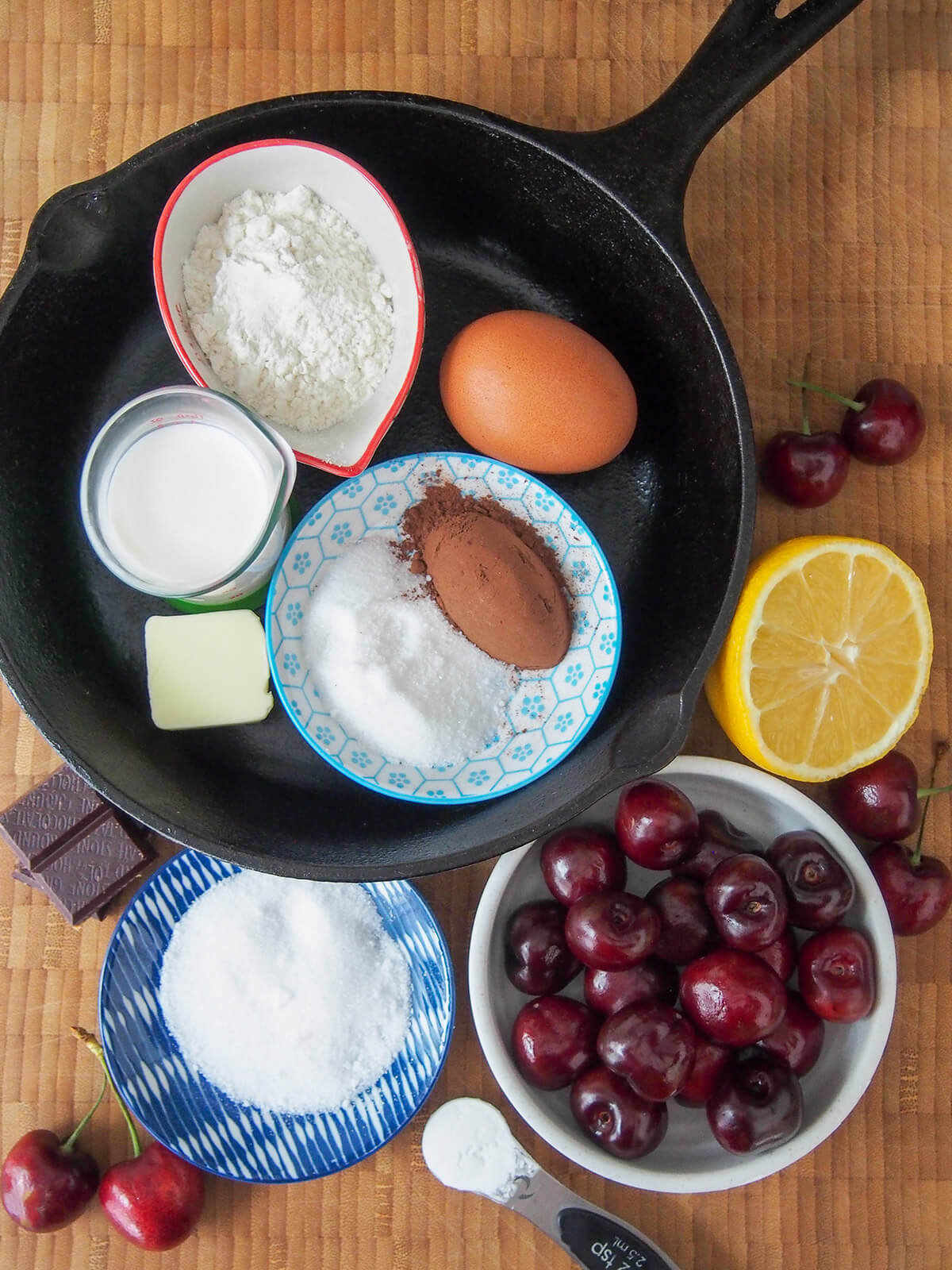 skillet with dishes of sugar, flour, cocoa powder and milk plus egg and piece of butter with lemon, cherries and sugar to side