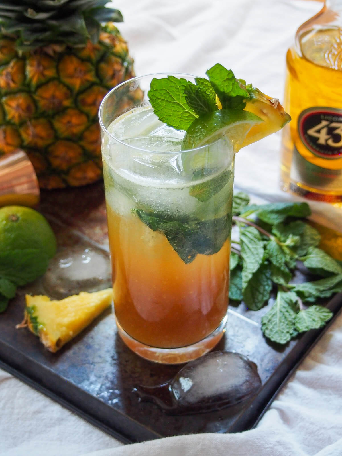glass of 43 pineapple mojito with pineapple and licor 43 bottle behind glass and ice cube in front