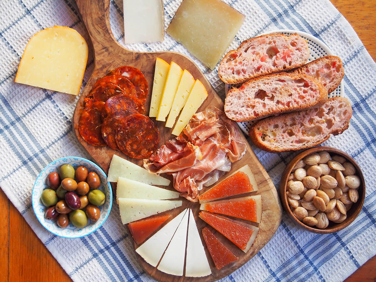 Spanish cheese board with ham and chorizo with plate of tomato bread and almonds to one side, olives other