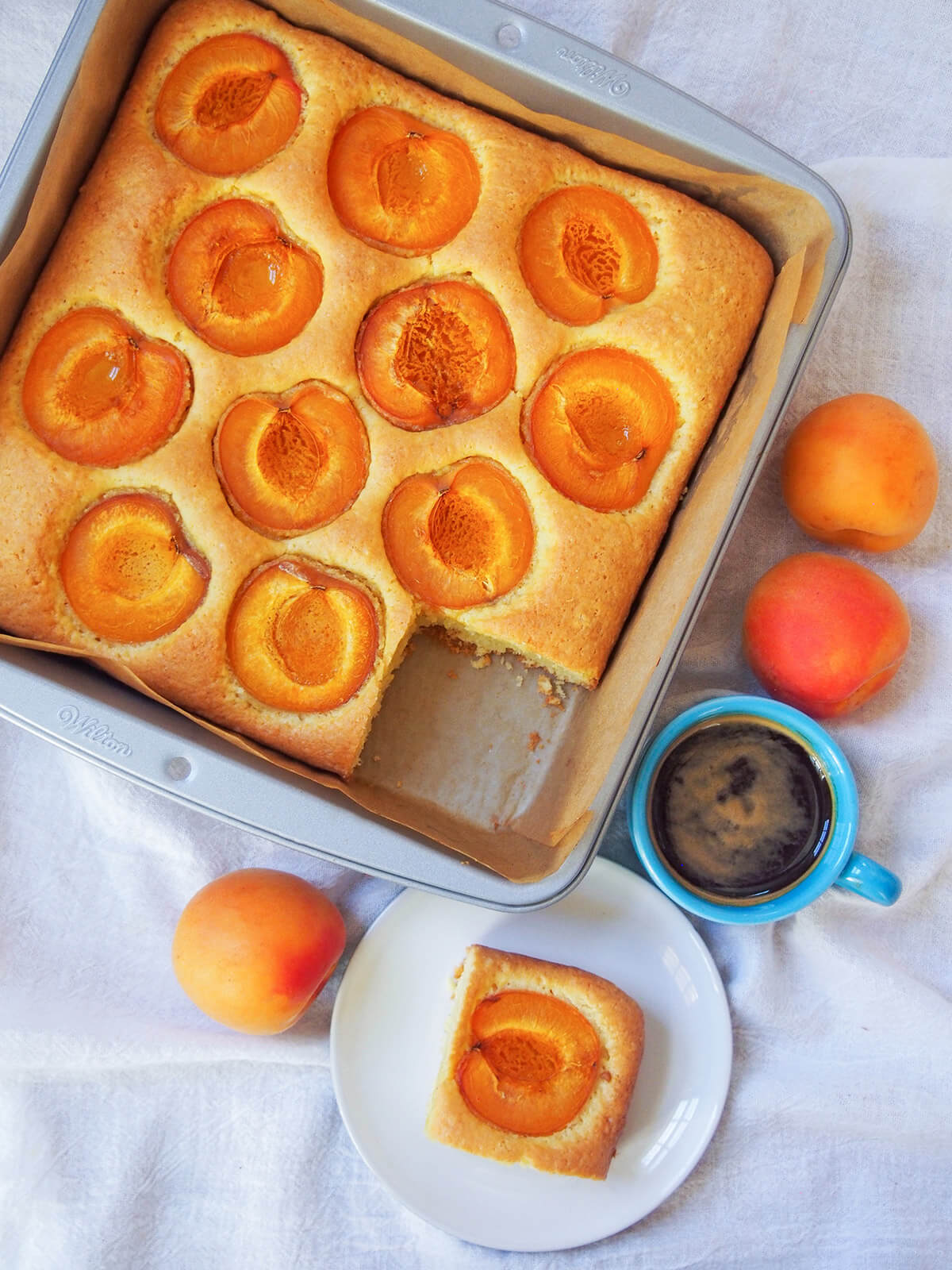 Austrian apricot cake is a wonderful combination of a simple cake base, with a touch of lemon, with large pieces of tender apricots nestled on top. It's a popular choice during the fruit's season alongside coffee, and makes a delicious anytime treat. apricot cake marillenkuchen in tin from overhead with slice on plate below