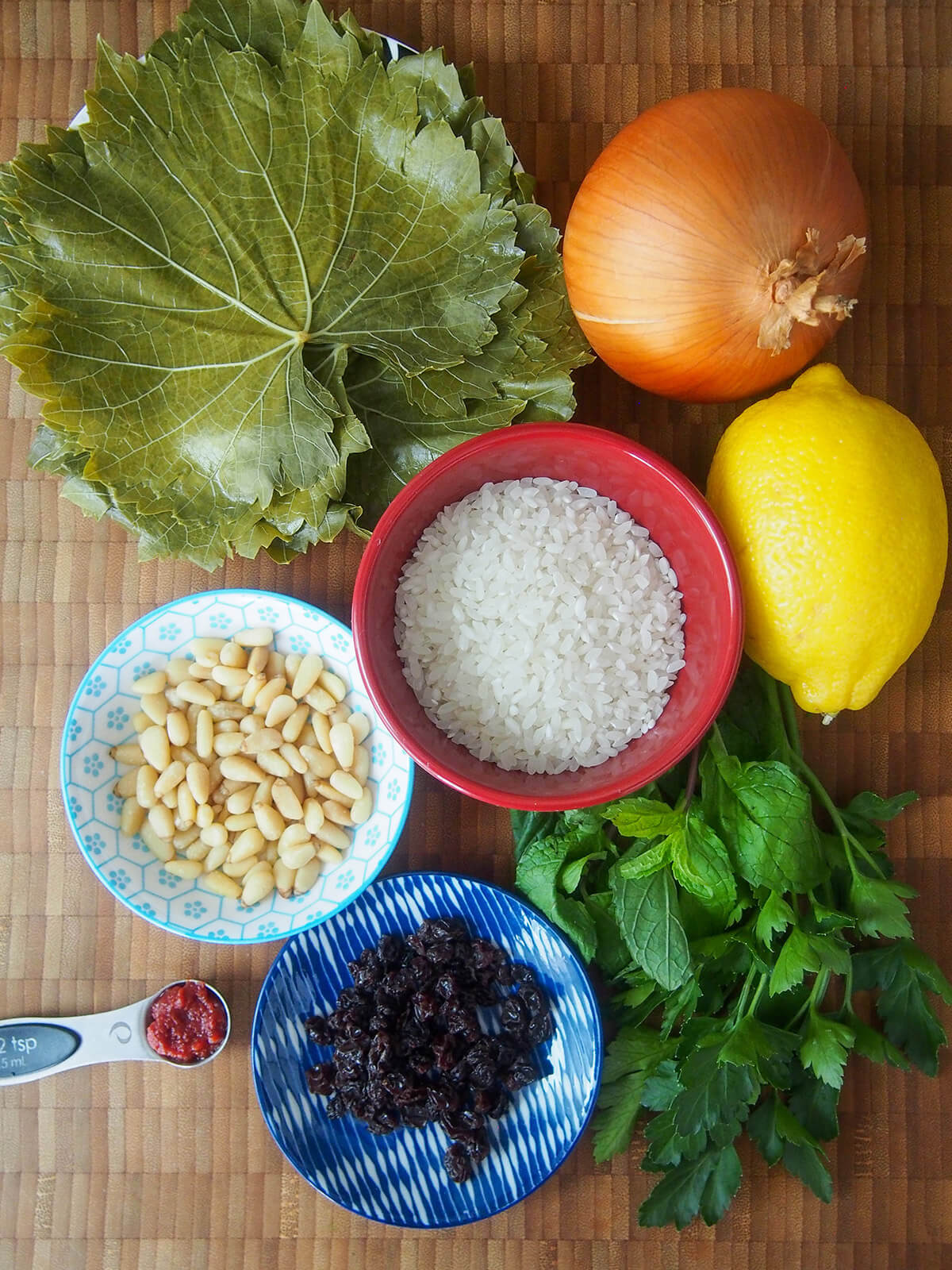 stack of grape leaves, bowl of rice, dishes with currants and pine nuts, and lemon, herbs and onion to side