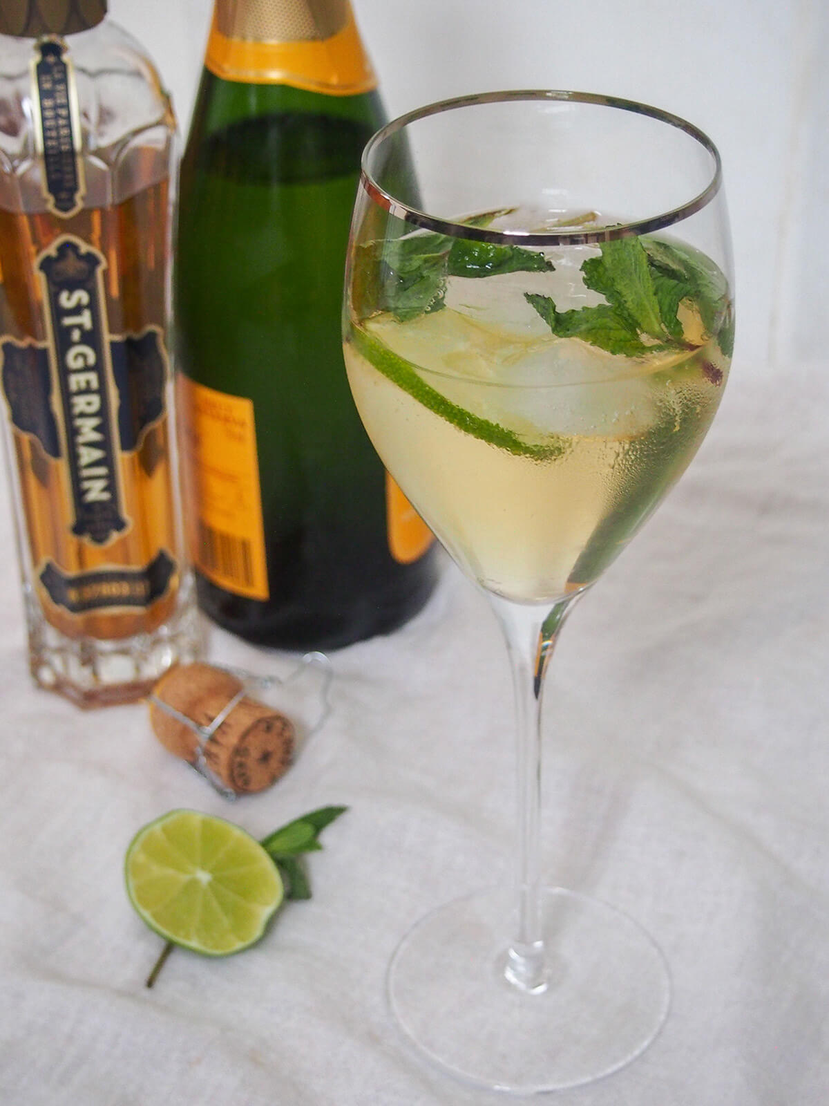 This Hugo spritz cocktail is easy to make, with a bright, refreshing flavor. It has a touch of aromatic sweetness from the St Germain, gentle bubbles and is nicely light. Perfect summer sipping. glass of Hugo spritz cocktail with bottles of prosecco and st Germain behind