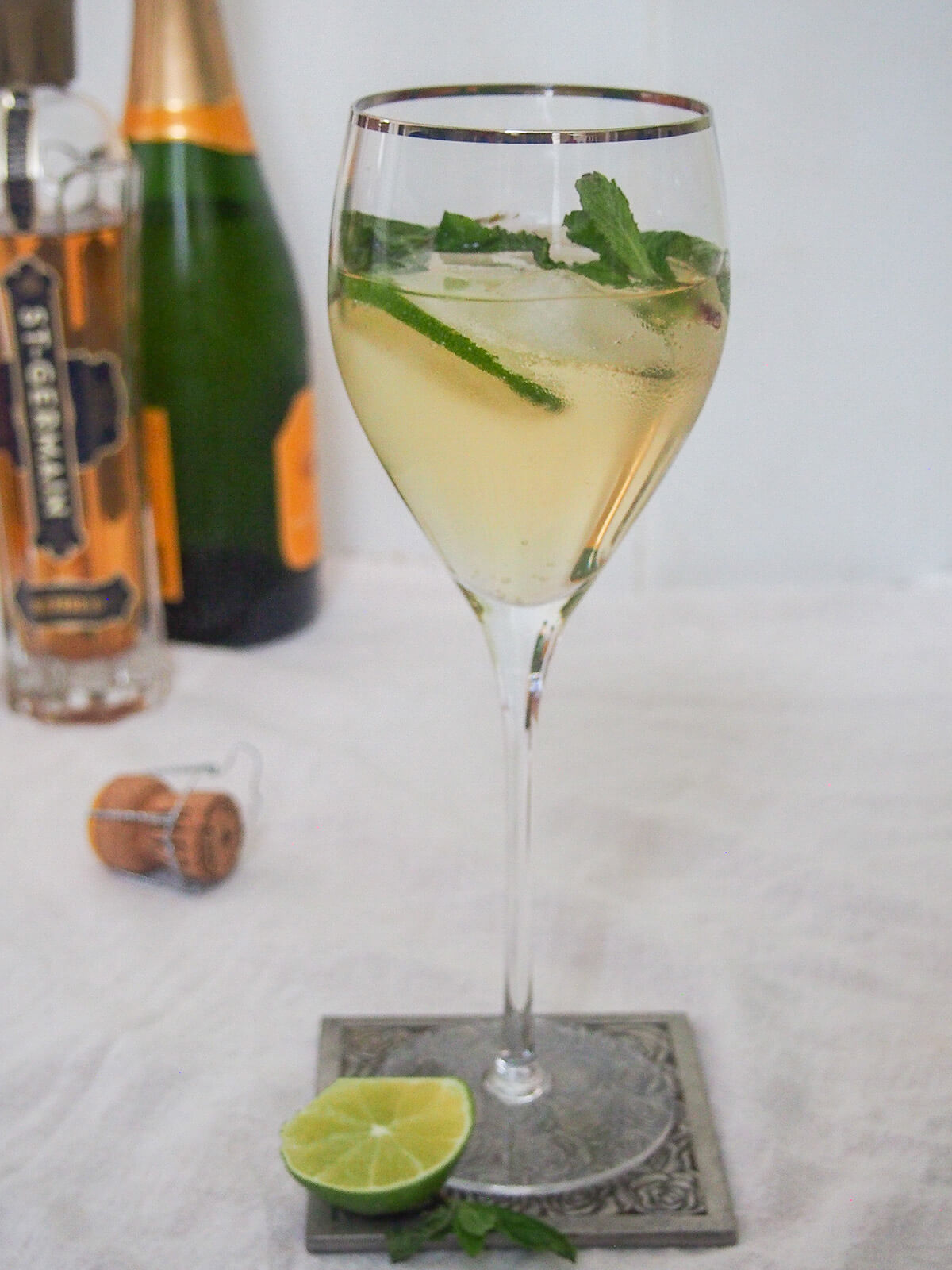 This Hugo spritz cocktail is easy to make, with a bright, refreshing flavor. It has a touch of aromatic sweetness from the St Germain, gentle bubbles and is nicely light. Perfect summer sipping. side view of glass of Hugo spritz cocktail with bottles in background