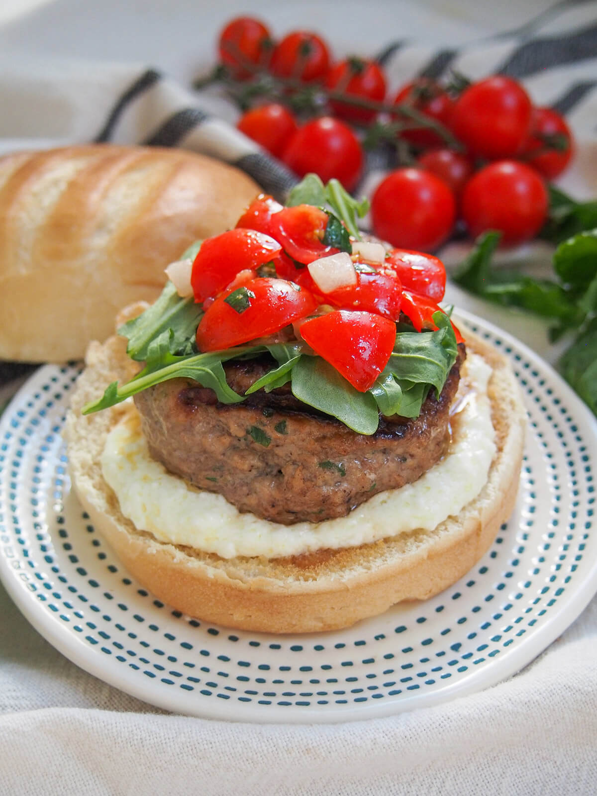 These lamb burgers are filled with Greek flavors, though they are presented in a less-than traditional way. Easy to make and incredibly tasty! lamb burger with feta sauce with vine of tomatoes behind