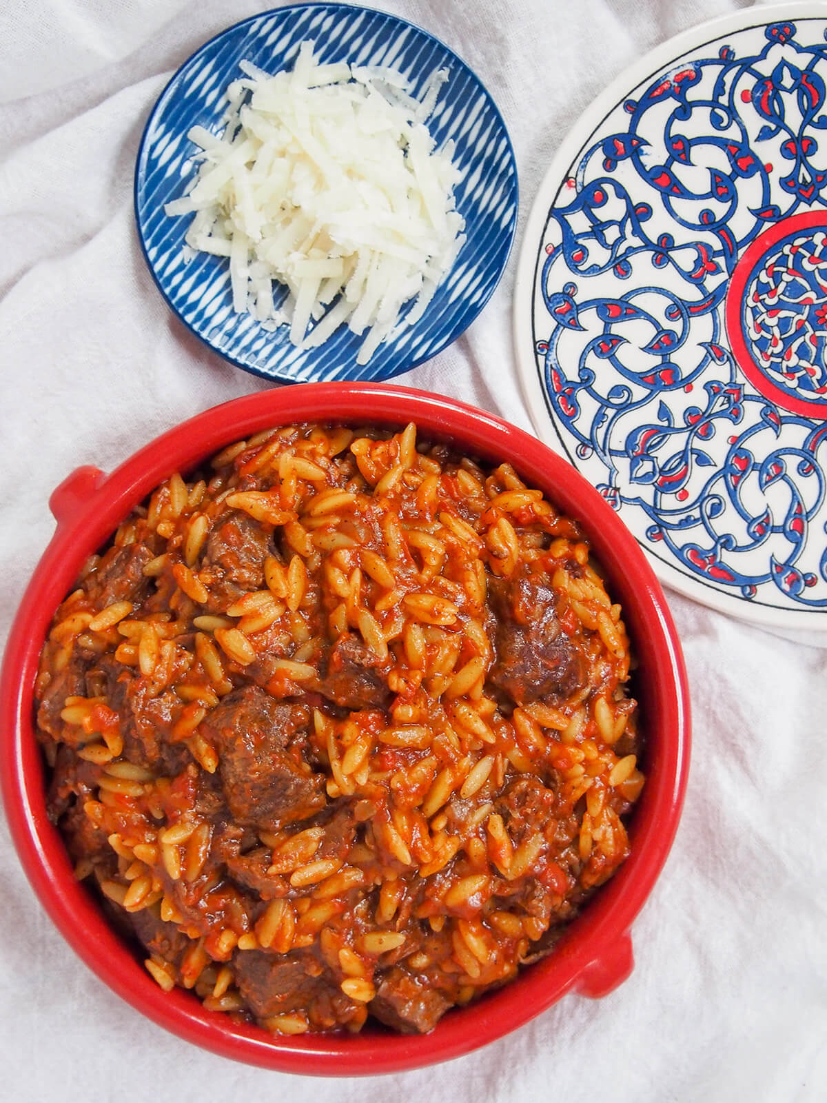 dish of youvetsi with small dish with cheese above and colored tile to side