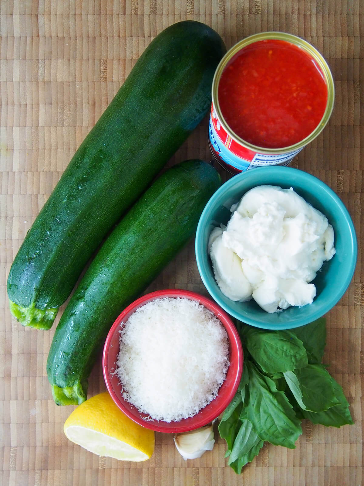 two zucchini next to bowls with ricotta, parmesan and can of tomato, basil, lemon