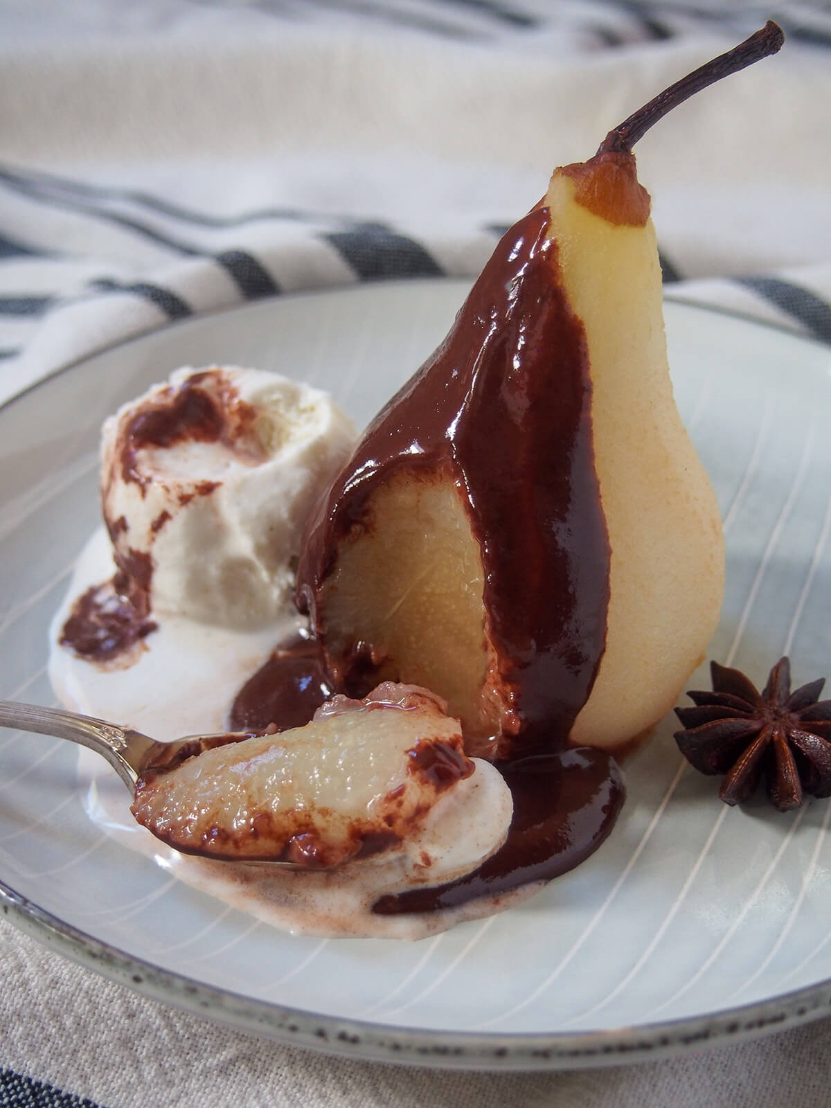 spoonful of pear sitting on plate in front of rest of poached pear with chocolate sauce aka poire belle Helene