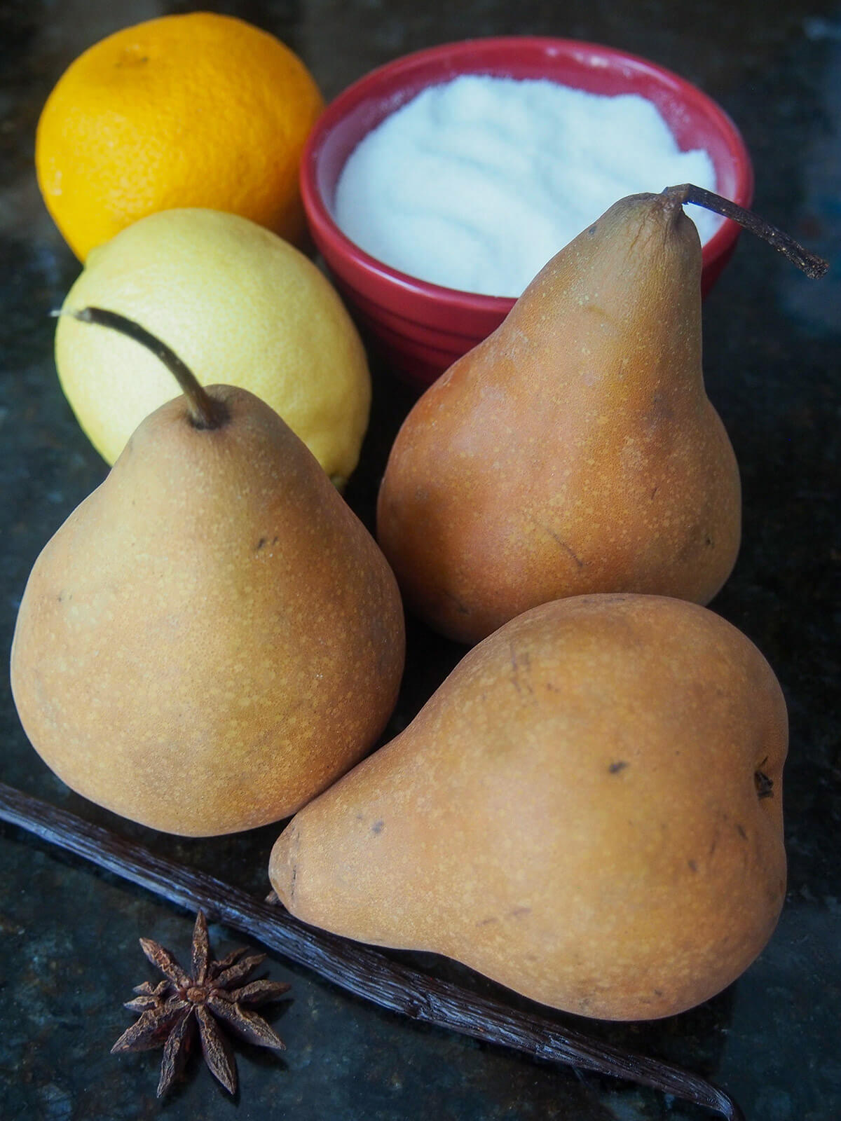 three pears, orange, lemon, bowl of sugar, vanilla pod and star anise on table.