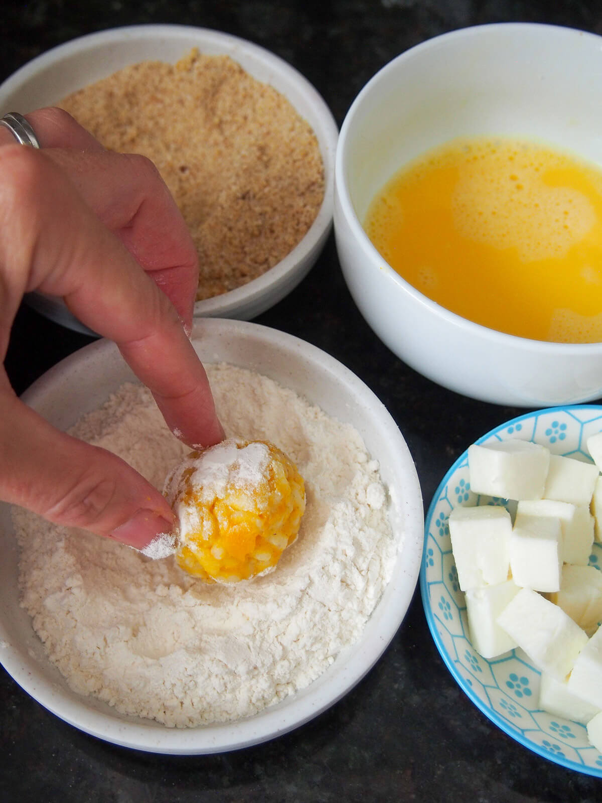 rolling arancini in flour with dishes of egg, breadcrumbs and mozzarella cubes to side
