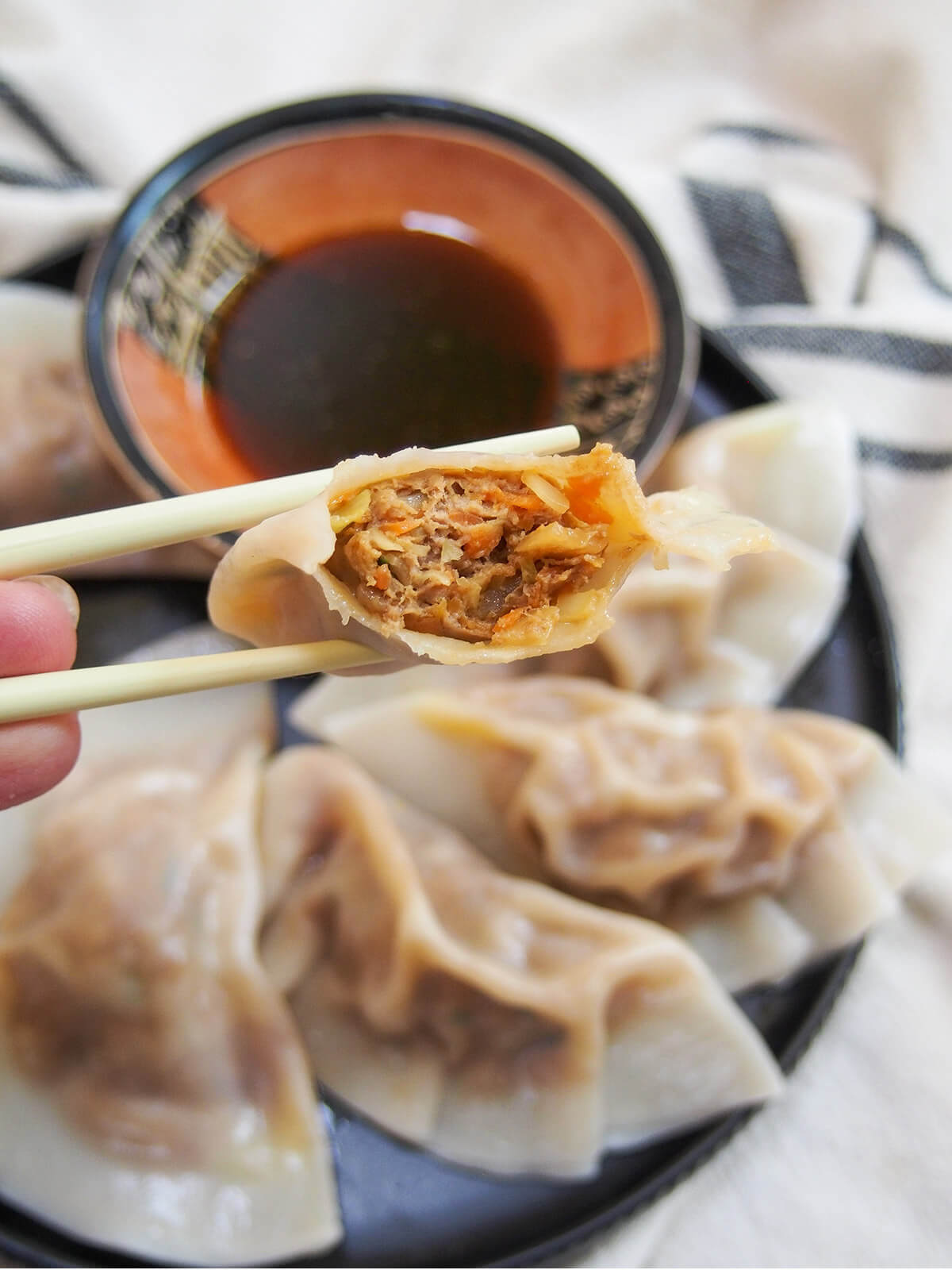 holding a bitten cumin lamb dumpling with chopsticks over plate of more