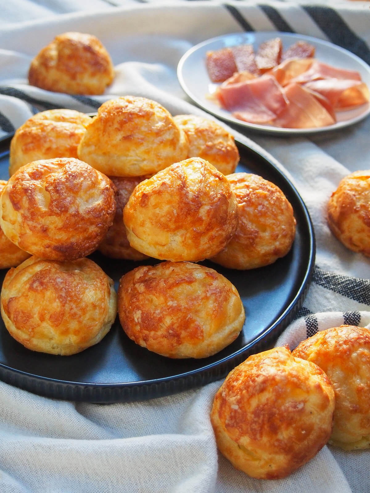 Gougères are classic French cheese puffs that make a great snack or canapé. Basically a savory choux pastry, with cheese added in, baked to crisp, light deliciousness. plate of gougeres cheese puffs from side with plate of ham behind