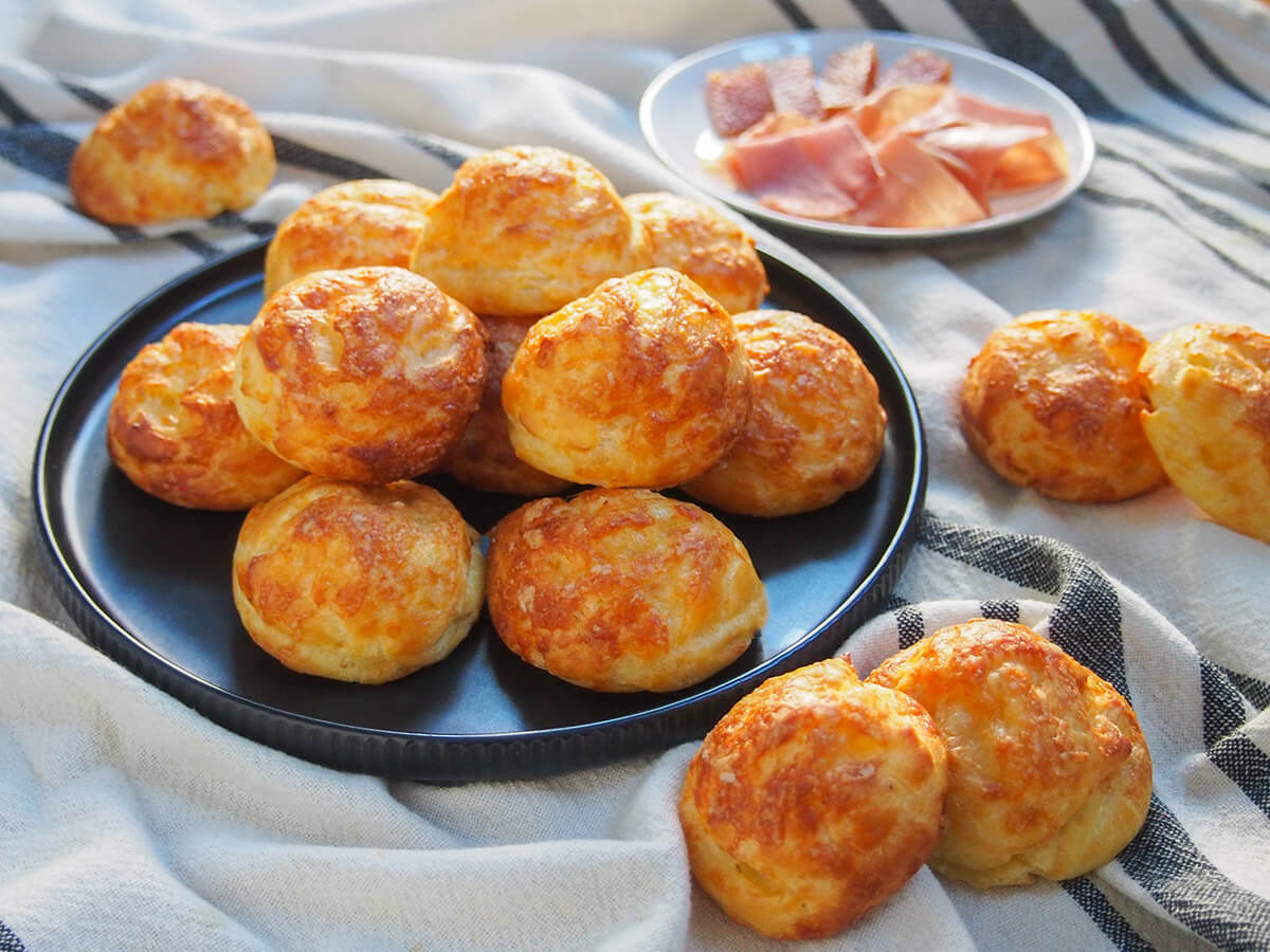Gougères are classic French cheese puffs that make a great snack or canapé. Basically a savory choux pastry, with cheese added in, baked to crisp, light deliciousness. plate of gougeres cheese puffs with some in front and small plate of ham behind
