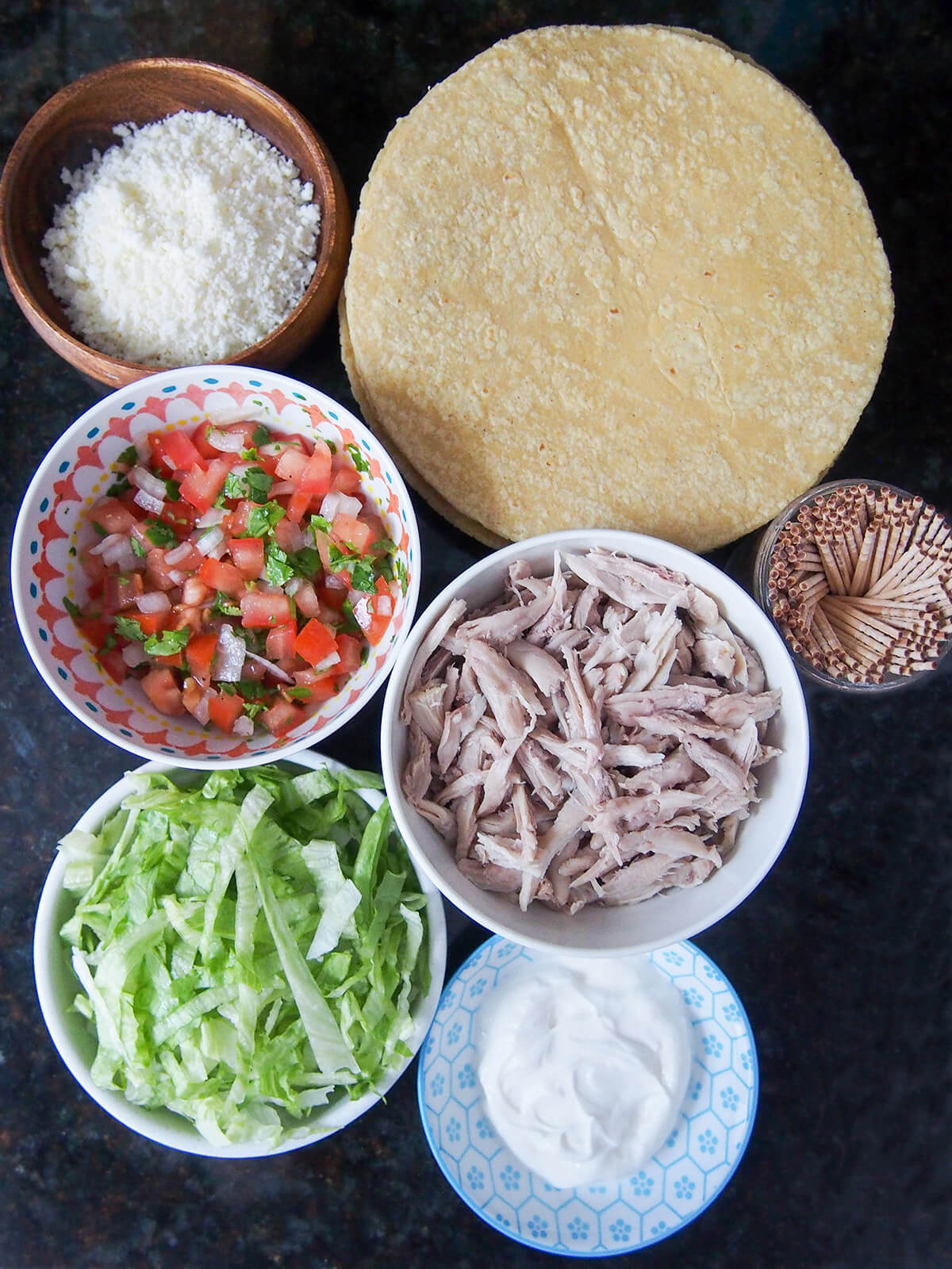 stack of corn tortillas, bowls of shredded chicken, pico de gallo, lettuce and cheese
