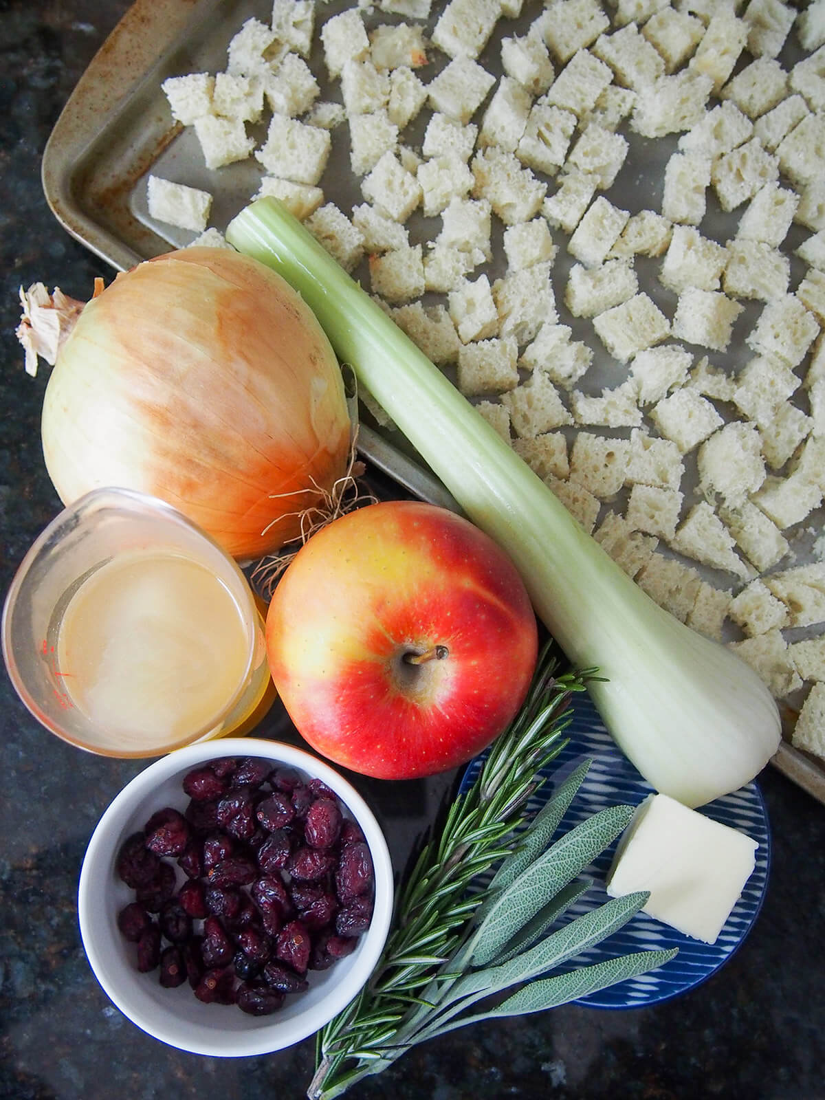 apple, onion, celery between baking sheet with bread cubes and dishes with cranberries, butter, saga and rosemary