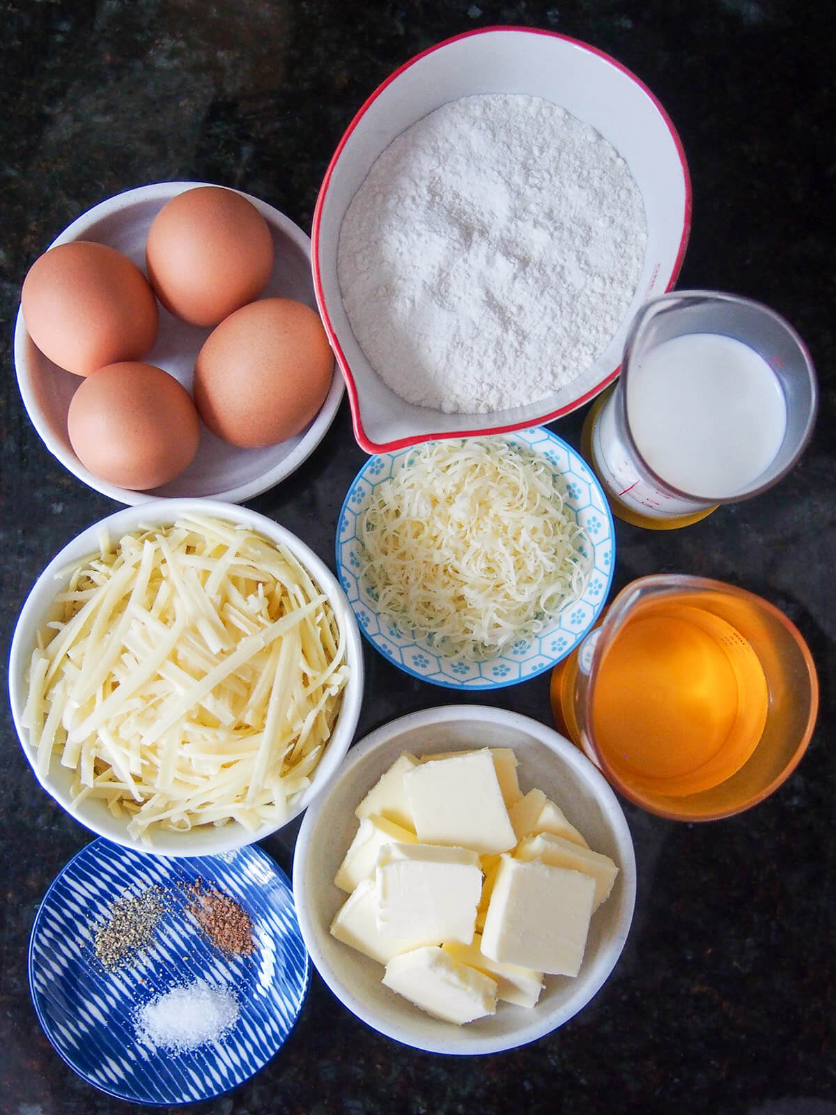 ingredients for gougeres bowls of flour, eggs, grated cheese, butter and measures with milk and water