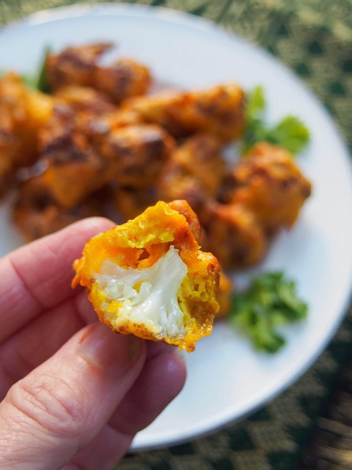 holding a bite of Cauliflower pakora, a tasty Indian appetizer holding a bitten into cauliflower pakora in hand over plate
