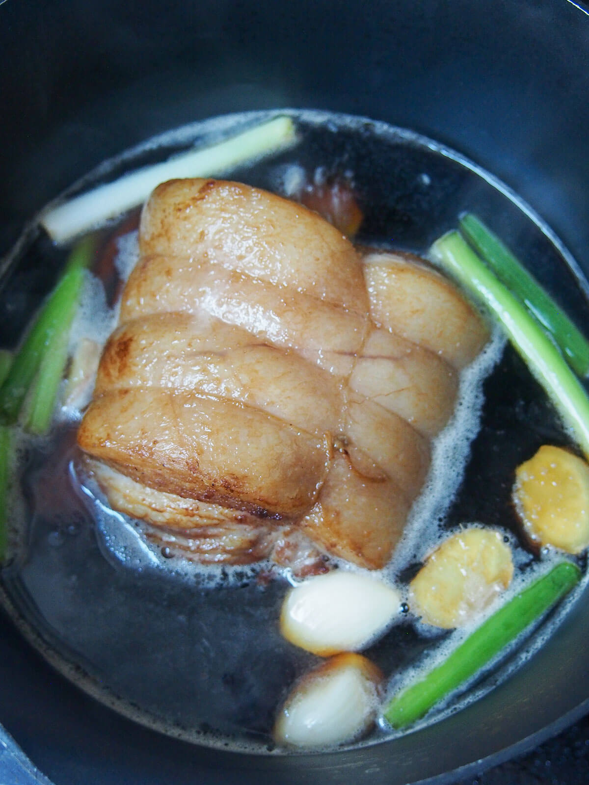 pork in braising liquid with pieces or ginger, garlic and green onion around pork